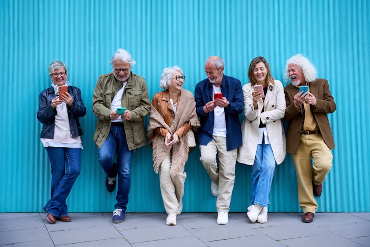 Group of older people standing against a bright blue wall, looking at their mobile phones and laughing.
