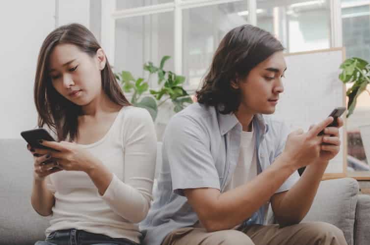 An Asian woman and South Asian man sit on a couch and use their smartphones