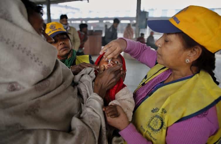 A volunteer wearing a vest and hat squeezes drops from a bottle into the mouth of a child, being held by its guardian.