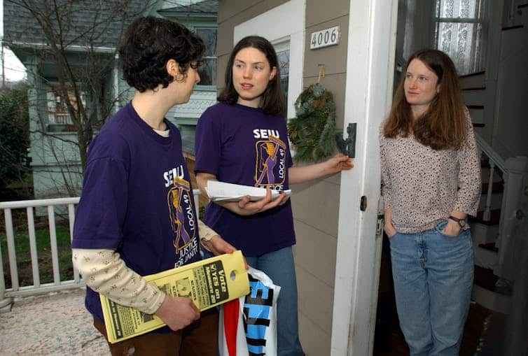 Two canvassers standing on the porch at the door with a resident of a home.
