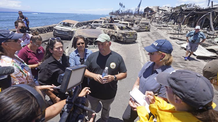 A man wearing a T-shirt with the state seal of Hawaii speaks with reporters, standing next to a woman with 'FEMA' on her cap and shirt with ocean and burned properties behind them.