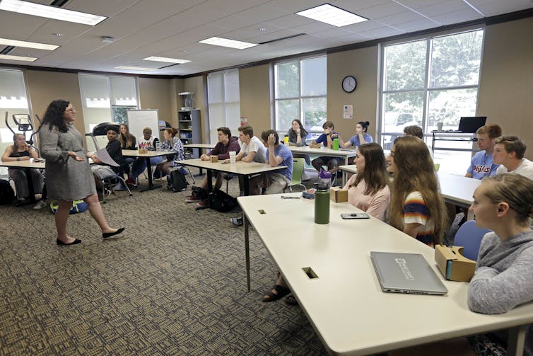 A sunny classroom full of students at long tables, with a female teacher in a dress pacing in front as she talks.