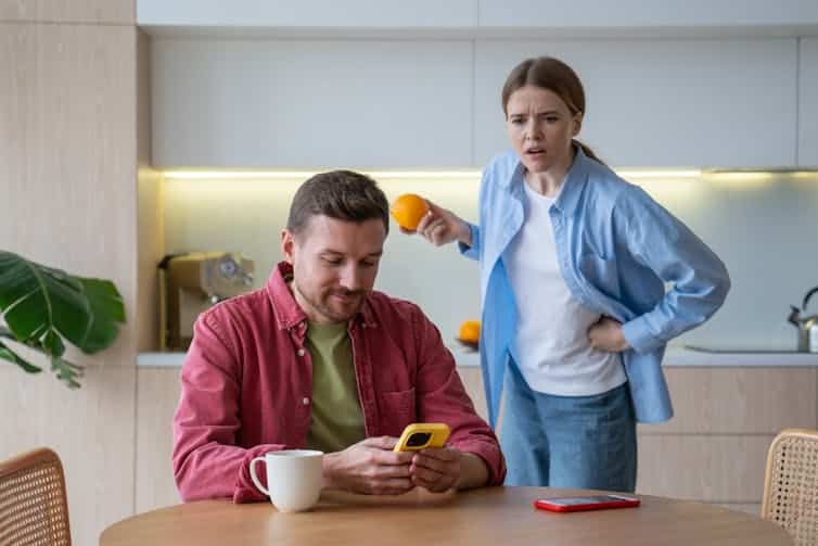 A man sits at a kitchen table looking at a smartphone. A woman behind him looks over his shoulder with an upset expression