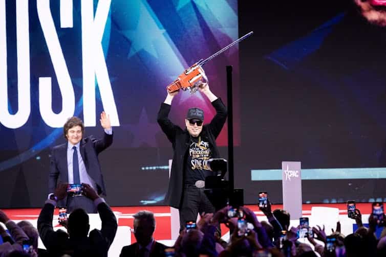 Musk, wearing a black Maga baseball cap and sunglasses, hoists a chainsaw over his head on stage, while Javier Milei waves behind him