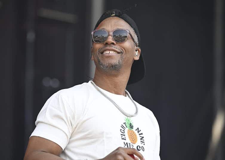 A man with sunglasses, a black baseball cap and white t-shirt smiles on a stage.